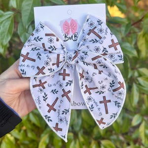 May include: White fabric bow with a pattern of brown crosses, floral accents, and the text "He is risen." The bow is held against a backdrop of green foliage. The bow has a decorative design.