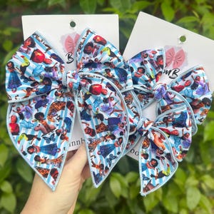 May include: Two large hair bows with a blue and white striped background and a colorful character print. The bows are attached to white cards with a pink bow logo. The bows are held up by a hand.