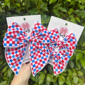 May include: Two hair bows with a red, white, and blue checkered pattern. The bows are attached to white cardstock with a logo. The bows are a patriotic accessory.