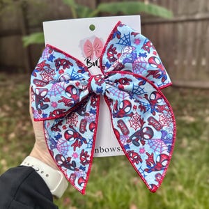 May include: A large, light blue hair bow featuring a repeating pattern of Spider-Man characters and webs. The bow has red trim and is attached to a white card with the text "Bows" and a logo.