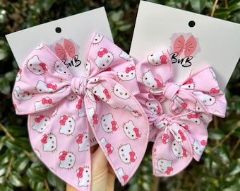Może przedstawiać: Dwie jasnoróżowe ozdoby do włosów z powtarzającym się wzorem Hello Kitty z białymi twarzami i czerwonymi kokardkami. Każdy element jest przymocowany do białej karty z otworem i czarnym napisem "BuB" oraz ikoną różowej kokardki.