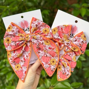 May include: Two hair bows attached to white display cards, featuring a light pink fabric with a repeating pattern of orange pumpkins, yellow sunflowers, and small white flowers. Each bow has a bright orange fabric trim along the edges.