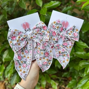 Op de afbeelding: Twee haarstrikken van bloemenstof met witte rand worden gepresenteerd op witte kaarten. De strikken hebben een patroon van roze rozen, gele bloemen en groene bladeren. De kaarten hebben het logo "BuB" in roze.