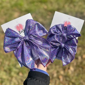 May include: Two purple hair bows with a shimmering holographic finish attached to white display cards. The larger bow is on the left and the smaller bow is on the right. Each card features a small pink bow icon and the text BuB printed in black.