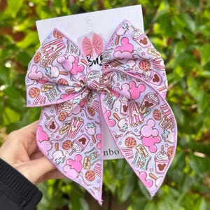 May include: A large hair bow featuring a light purple background with a repeating pattern of pink mouse-shaped balloons, popcorn buckets, cookies, and drinks. The fabric has a glossy finish and is attached to a white display card with a logo.