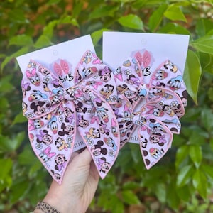 May include: Two fabric hair bows featuring a light pink background with a repeating pattern of cartoon characters including Minnie Mouse, Daisy Duck, and Pluto, accented by small white daisies and finished with a pink stitched edge on white display cards.