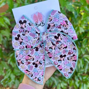 May include: A large hair bow with a repeating pattern of Mickey and Minnie Mouse characters, ice cream, and other themed elements on a light pink background. The bow has a light blue trim and is attached to a white card.