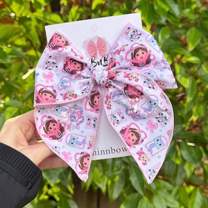 May include: A large hair bow featuring a white fabric with a repeating pattern of cartoon characters, cats, and pink bows. The bow is attached to a white display card with the text BuB and ninnbow printed on it, showcasing a pink bow logo.