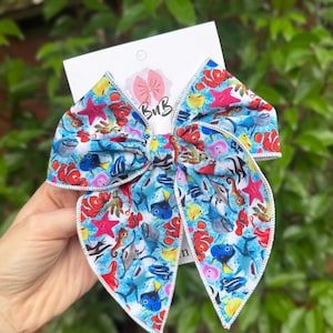 May include: A large, fabric hair bow featuring a colorful underwater scene with fish, starfish, and sharks on a blue background. The bow has a white stitched edge and a small tag with the letters "BvB".