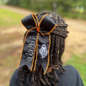 May include: A black hair bow featuring orange zigzag stitched edges, an embroidered white ghost graphic, and the name Alayah stitched in white cursive script. The ribbon is tied in a classic bow shape with two long, pointed tails hanging down.