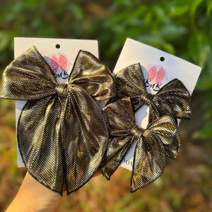 Puede incluir: Tres lazos para el cabello con un acabado texturizado en dorado metálico y negro, sujetos a tarjetas de presentación blancas. El lazo más grande está a la izquierda, mientras que dos más pequeños están a la derecha. Cada tarjeta tiene un logo BuB.