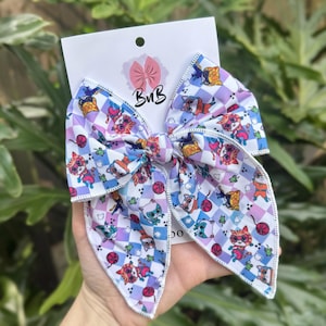 May include: A large fabric hair bow featuring a repeating pattern of cartoon cats in superhero masks and costumes over a light purple and white checkered background. The bow is attached to a white display card with the text BvB and a bow icon.