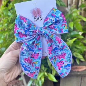 May include: A large, colorful hair bow featuring a repeating pattern of cartoon ponies in various colors on a light blue background. The bow has a lavender trim and is attached to a white card with a logo that reads "BnB".