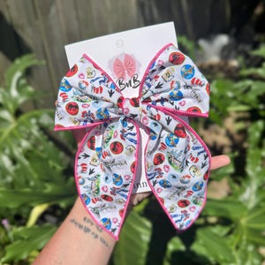 May include: A white bow with a pink trim and a colorful print of various images, including a cat in a hat, a red and white striped hat, and a pair of glasses. The bow is tied with a knot in the center and has two long tails.