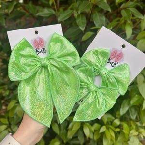 May include: Two bright green hair bows with a shimmering finish are displayed against a backdrop of green foliage. The bows are attached to white cards with the text "BuB" and a pink flower logo.