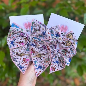 May include: Two large, light pink hair bows with a cartoon character print and pink trim. Each bow is attached to a white card with a smaller pink bow. The bows are held up by a person.
