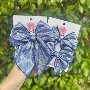 May include: Three blue hair bows with a shimmering, iridescent finish. The bows are attached to white cards with a pink logo that says "BuB". The bows are held in front of a green leafy background.
