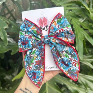 May include: A large, colorful hair bow featuring a whimsical print with red trim. The bow is made of fabric with a vibrant pattern of cartoon characters and scenes. The bow is attached to a white card with a logo.