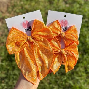 May include: Three bright orange hair bows with a shimmering, glittery texture attached to white display cards. The largest bow is on the left, while two smaller bows are stacked on the right. Each card features a small pink bow logo and the text BuB.