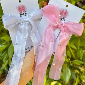 May include: Two hair bows made of sheer, translucent fabric, one white and one light pink. Each bow features long, flowing ribbons embellished with small, round, white faux pearls scattered across the surface. The bows are attached to white display cards.