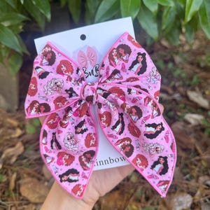 May include: A large pink hair bow featuring a repeating pattern of diverse illustrated figures with various hairstyles, small pink high-heeled shoes, and tiny white stars. The bow is attached to a white display card with a logo and text.