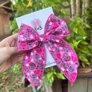 May include: A bright pink hair bow with a repeating pattern of cartoon characters, hearts, and other graphics. The bow is tied in a classic style with long tails. The bow is attached to a white card with the text "BuB" and "nnbow".
