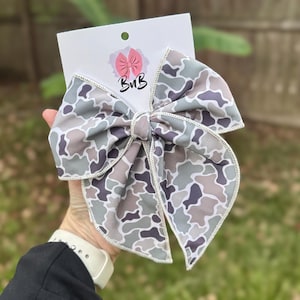 May include: A large hair bow with a camouflage pattern in shades of grey, brown, and purple. The bow is attached to a white card with a pink logo and the letters "BnB". The bow is held in a person's hand.
