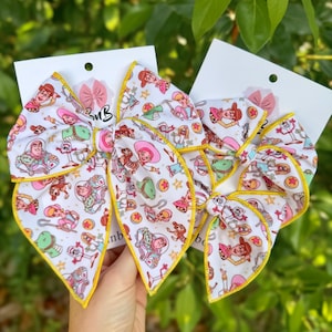 May include: Two white fabric hair bows featuring a repeating pattern of cartoon characters including a cowboy, a cowgirl, a space ranger, and a plastic fork toy, accented with yellow stitched edges and mounted on white display cards.