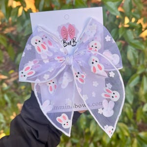 Op de afbeelding: Een grote, lichtpaarse haarstrik met een doorschijnende overlay met witte konijntjes met roze accenten en kleine witte bloemen. De strik is in het midden geknoopt en heeft een witte rand. De tekst "mmminabows.com" is zichtbaar.