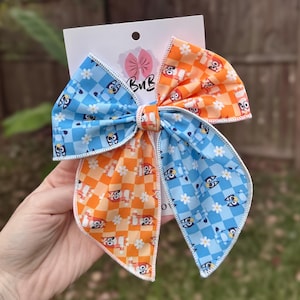 Könnte beinhalten: Eine große, handgefertigte Haarschleife mit geteiltem Design. Die blaue Seite zeigt ein Cartoon-Charakter- und weißes Blumenmuster, während die orangefarbene Seite ein passendes Cartoon-Charakter- und weißes Blumenmuster aufweist. Die Schleife ist an einer weißen Karte befestigt.
