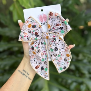 Puede incluir: Un lazo para el pelo grande, hecho a mano, con un fondo blanco y un patrón repetido de personajes y símbolos de Star Wars. El lazo tiene un borde dorado y está sujeto a una tarjeta blanca con el logotipo "BuB".