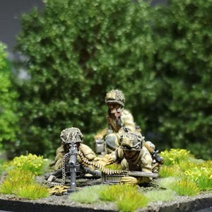 Może przedstawiać: Miniaturowe figurki żołnierzy w kamuflażowych mundurach, ustawione w trawiastym otoczeniu z karabinem maszynowym. Figurki są szczegółowe z hełmami i wyposażeniem, na tle zielonych liści.