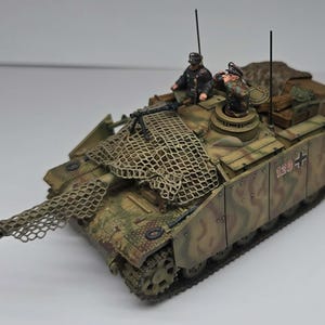 Op de afbeelding: Gedetailleerd model van een militaire tank, geschilderd in camouflagekleuren bruin, groen en beige. De tank heeft een lange loop, een netbedekking en twee miniatuurfiguren. Het nummer "120" is zichtbaar aan de zijkant.