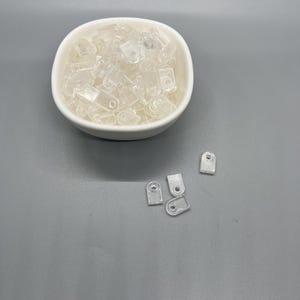 May include: A white bowl filled with clear plastic curtain rings. Five additional rings are scattered on a grey surface.
