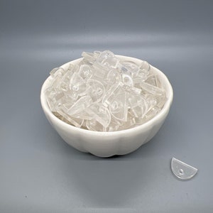 May include: A white ceramic bowl filled with clear plastic half-circle shapes. A single clear plastic half-circle shape is on the grey surface next to the bowl.