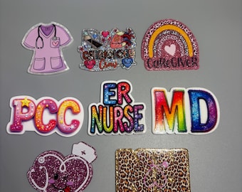 May include: A collection of colorful medical-themed badge reels. Designs include a pink scrub top, a rainbow with a heart, and text that reads "ER NURSE" and "MD". Other designs feature a heart with a stethoscope and a leopard print stethoscope.