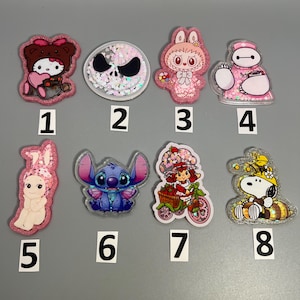 Könnte beinhalten: Eine Sammlung von acht farbenfrohen, glitzernden Charakter-Anhängern. Die Anhänger zeigen Designs von Hello Kitty, Jack Skellington, einem rosa Hasen, Baymax, einer hasenartigen Figur, Stitch, Erdbeerkuchen und Snoopy.