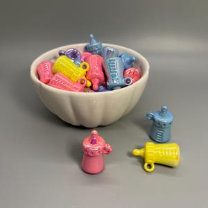 May include: A white bowl filled with colorful baby bottle charms in shades of pink, yellow, blue, and purple. Several charms are scattered outside the bowl. The charms have a glossy finish and are designed with a handle and a nipple.