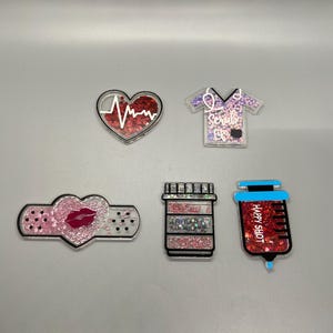 Puede incluir: Cinco dijes de resina transparente con brillo y texto. Los dijes tienen forma de corazón con una línea de latido del corazón, una bata de trabajo con el texto "Scrub Life", una venda con un labio rojo, una botella de píldoras con el texto "You're My Medicine", y una jeringa con el texto "Happy Shot".