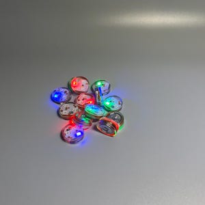 May include: A collection of small, round LED lights in various colors, including red, green, and blue. The lights are arranged in a pile, with some emitting a bright glow. These battery-operated lights are suitable for crafts and decorations.