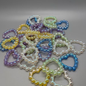 Puede incluir: Una colección de abalorios en forma de corazón iridiscentes hechos de cuentas de plástico. Los abalorios son de varios colores, incluyendo morado, azul, amarillo y verde.