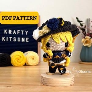 May include: Crocheted amigurumi doll with yellow hair, a black hat with a blue flower, and a black and white outfit. The doll is standing on a wooden base. The image includes the text "Krafty Kitsune" and "PDF Pattern".