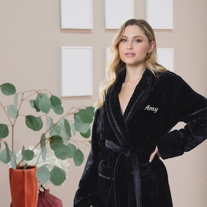 May include: A plush black robe featuring a shawl collar and a matching tie belt at the waist. The left side of the chest is embroidered with the name Amy in white cursive script. The robe has long sleeves and a soft, textured fabric finish.