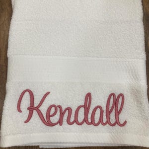 May include: White bath towel with the name "Kendall" embroidered in a cursive, rose-coloured font. The towel has a textured surface and a decorative band near the top. The towel is folded.
