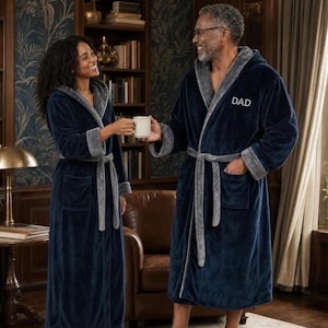 May include: Two plush, navy blue robes with contrasting grey shawl collars, cuffs, and waist ties. The robes feature long sleeves, front patch pockets, and a hood. The right robe has the word DAD embroidered in white on the left chest area.