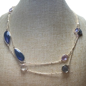 May include: A gold-toned necklace with multiple gemstones of various shapes and colors. The necklace features teardrop-shaped, oval, and round stones in shades of blue, purple, and clear. The necklace is displayed on a neutral-toned mannequin.