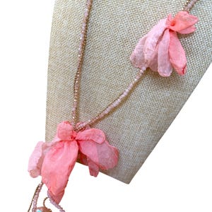 May include: A delicate necklace featuring a gold chain and strands of small, light pink beads. The necklace is adorned with several large, layered, light pink fabric flower pendants, creating a soft and feminine aesthetic.
