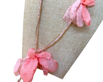May include: A delicate necklace featuring a gold chain and strands of small, light pink beads. The necklace is adorned with several large, layered, light pink fabric flower pendants, creating a soft and feminine aesthetic.