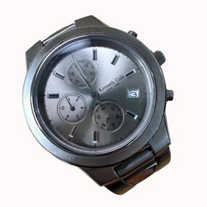 May include: A silver-toned Kenneth Cole New York wristwatch with a stainless steel band. The watch face has three sub-dials, rectangular hour markers, and a date window at the 3 o'clock position. The watch is water resistant.