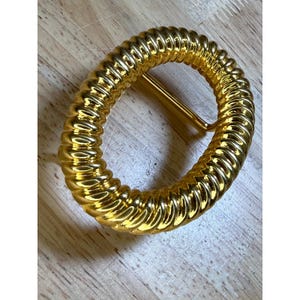 May include: Oval-shaped gold-colored belt buckle with a ridged design. The buckle has a gold-colored bar on one side for the belt to slide through. The buckle is made of metal and is on a wooden surface.